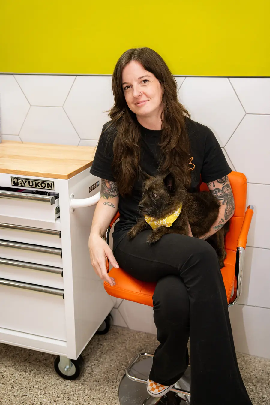 Ava, owner and groomer at Black Dog Grooming Salon, seated in the shop holding a small dog in a yellow bandana